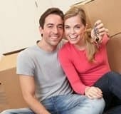 A happy couple sitting on the floor, holding house keys, surrounded by moving boxes in their new home, ready to begin storing their belongings.