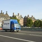 A blue self storage company truck driving over a bridge with city buildings in the background under a clear sky.