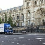 A blue delivery truck labeled "strong.com" drives past the ornate facade of the natural history museum in London, advertising self storage solutions.