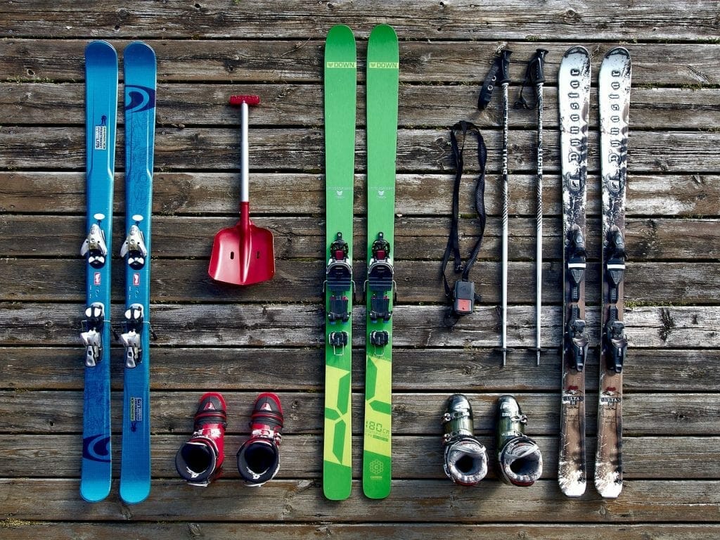 Various ski equipment, including blue and green skis, poles, boots, and a red shovel, arranged neatly on a wooden plank background for storing in self storage.