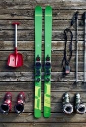 Various ski equipment, including blue and green skis, poles, boots, and a red shovel, arranged neatly on a wooden plank background for storing in self storage.
