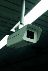 Security camera mounted on a ceiling with fluorescent lights in a dimly lit self storage room.