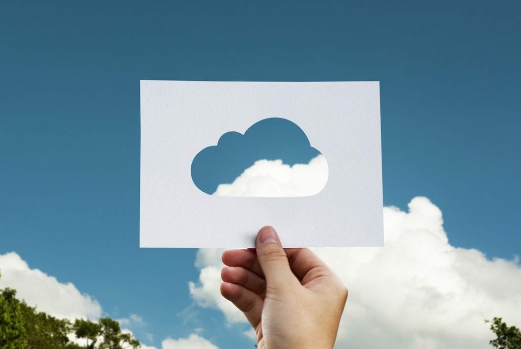 A hand holding up a piece of paper with a cloud-shaped cutout, aligning the cutout with a real cloud in a blue sky, symbolizing creative storing of visual elements.