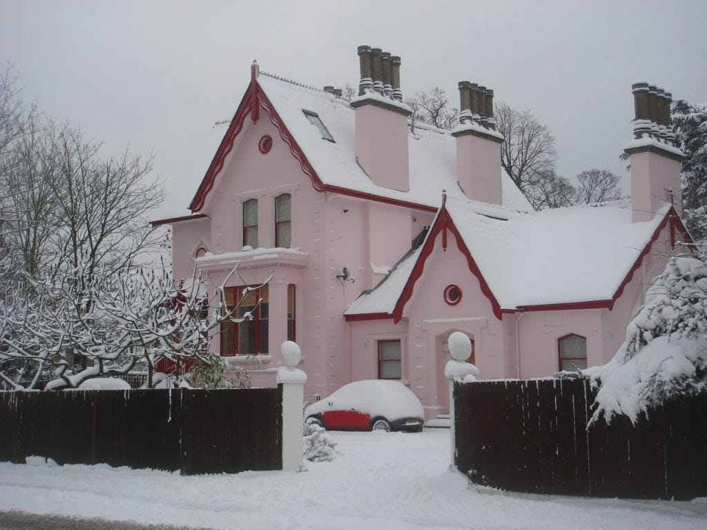 A pink house with multiple chimneys covered in snow, with a white car also dusted with snow in the driveway near the self storage unit.
