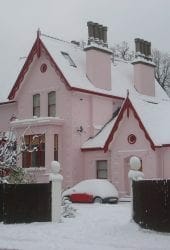 A pink house with multiple chimneys covered in snow, with a white car also dusted with snow in the driveway near the self storage unit.