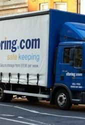 A blue delivery truck labeled "self storage.com" on a city street, with a red truck in the background.