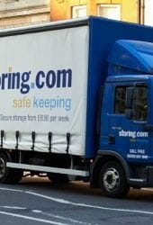 A blue delivery truck with the "storing.com" logo navigates a bustling city street, a building and red vehicle in the background. It's as seamless as moving home with a dog, ensuring everything flows with as little stress as possible.
