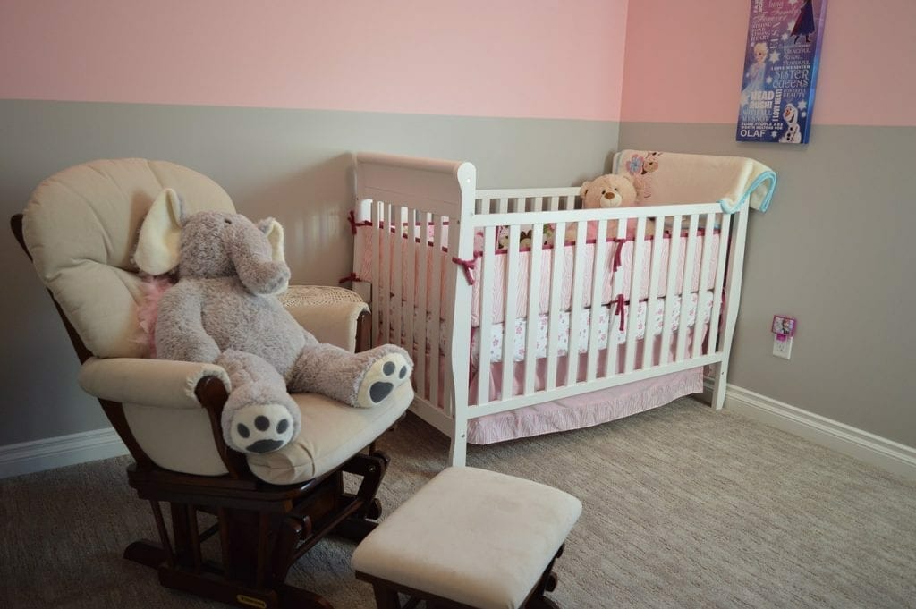 A cozy nursery with a white crib, a rocking chair equipped for storing with a large stuffed elephant, and pink walls.