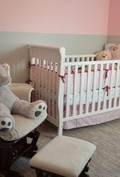 A cozy nursery with a white crib, a rocking chair equipped for storing with a large stuffed elephant, and pink walls.