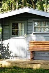 A small wooden cabin with a white door and a bench on the front porch, situated in a lush green forest, perfect for storing outdoor equipment.