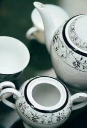 An elegant white porcelain tea set with black floral designs, perfect for storing hot beverages, including a teapot, lid off, and multiple cups on a dark table.