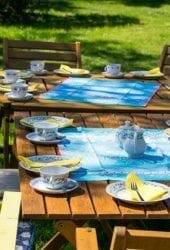 Outdoor wooden dining table set with teacups, plates, and a teapot, ready for a meal in a sunny garden, ideal for storing conveniently after use.