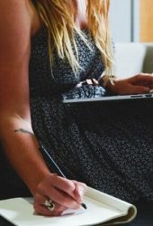 Woman in a dotted dress sitting on a couch, typing on a laptop and writing in a notebook, with a visible tattoo on her arm and self storage documents beside her.