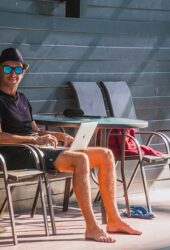 Man in sunglasses and hat using a laptop at a poolside table with empty chairs next to him, storing his belongings securely nearby.