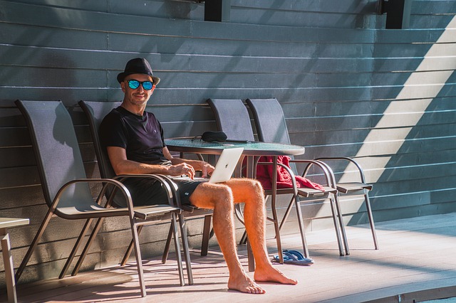 Man in sunglasses and hat using a laptop at a poolside table with empty chairs next to him, storing his belongings securely nearby.