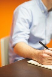 A man in a blue shirt storing notes on a pad while working on a laptop in an office with an orange wall.