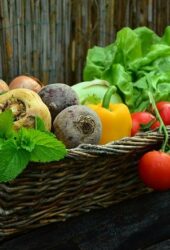 A wicker basket filled with fresh vegetables, including tomatoes, bell peppers, beets, and lettuce, set against a wooden and reed background ideal for storing produce.