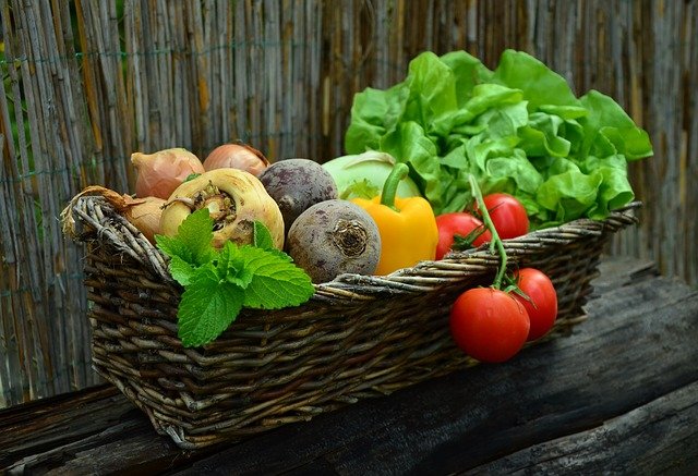 A wicker basket filled with fresh vegetables, including tomatoes, bell peppers, beets, and lettuce, set against a wooden and reed background ideal for storing produce.