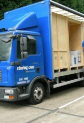 Two men in blue shirts load large wooden crates into a blue delivery truck on a paved road, surrounded by trees, perhaps destined for cheap storage units nearby.