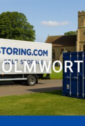 Moving truck and blue storage container sit on a road beside a green lawn with a stone church and trees in the background.