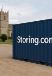 Two blue shipping containers with "Storing.com" are on a dirt road near a church with a tall tower and trees in the background.
