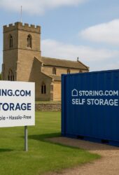A blue storage container and a sign reading "Storing.com Self Storage" are positioned on a grassy area in front of a stone church building.