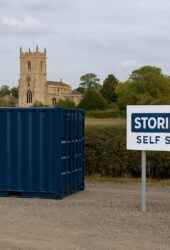 A blue shipping container sits next to a sign reading "STORING.COM SELF STORAGE" in front of a hedge. A church with a tower is visible in the background.