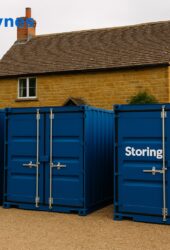 Three blue storage containers labeled "Storing.com" are placed outside a traditional stone house with trees in the background. The text "Milton Keynes" is in the top left corner.